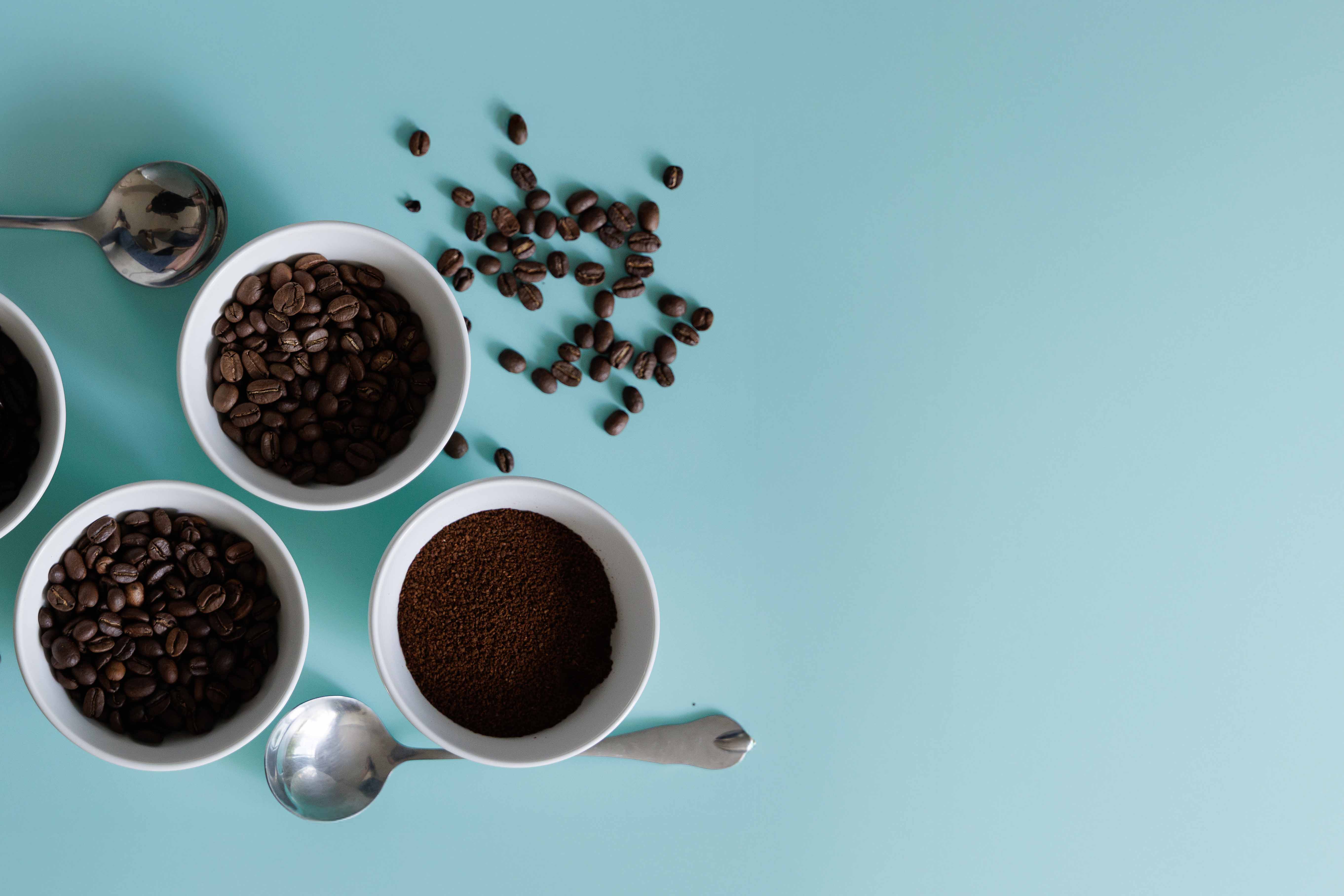 Three mugs with coffee beans and grounds on a light blue background. Coffee beans are spilled in the background.