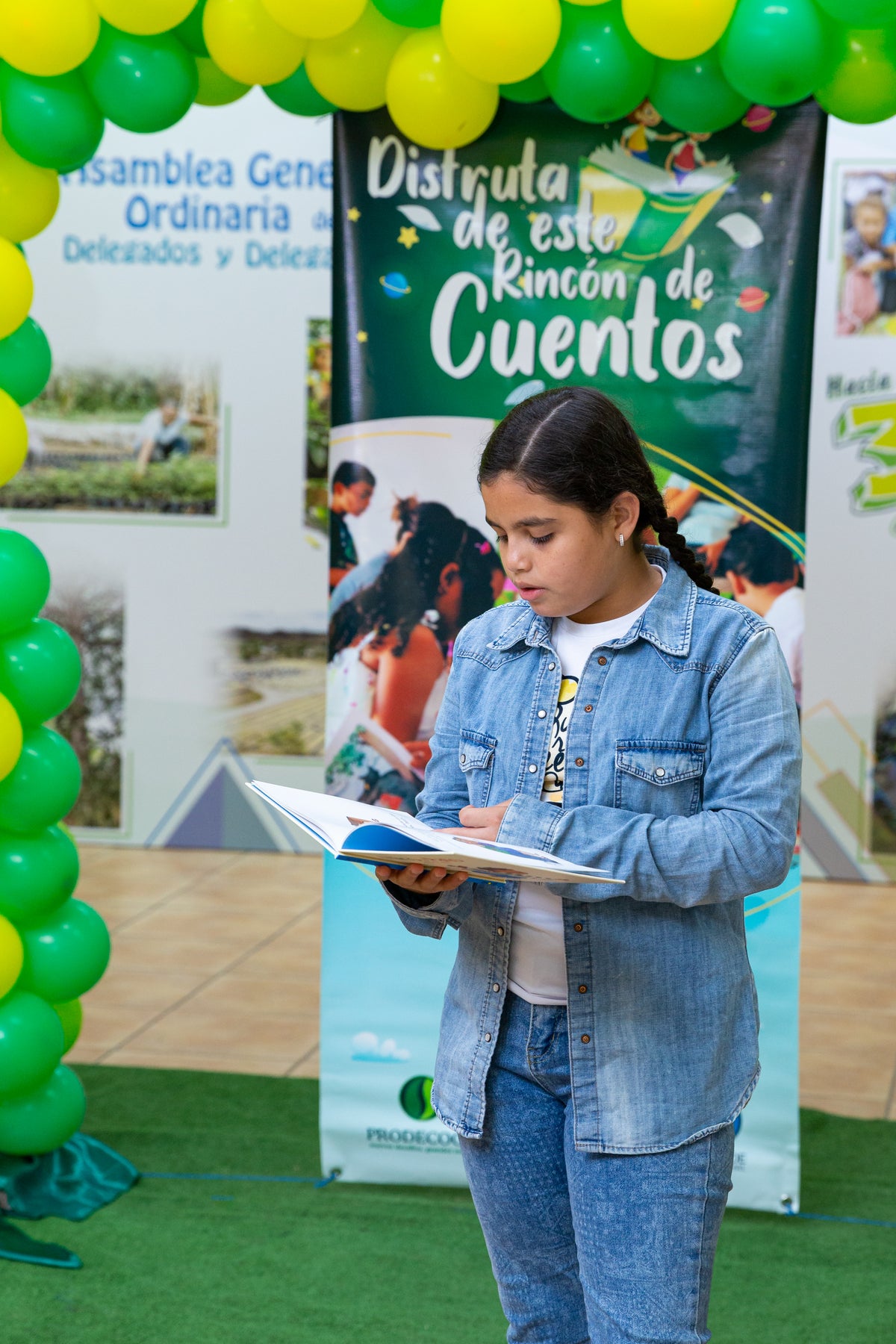 Young girl reading a book in front of a promotional banner with green and yellow balloons.