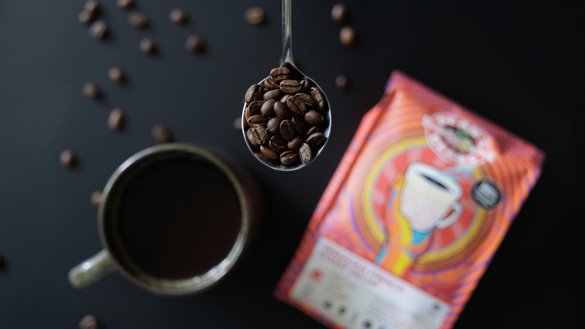 Spoonful of coffee beans over a cup of coffee with a colorful dean's beans coffee package on a dark surface.