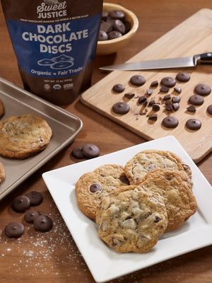 A bag of dark chocolate discs, some chocolate on a cutting board with a knife, and a plateful of chocolate cookies